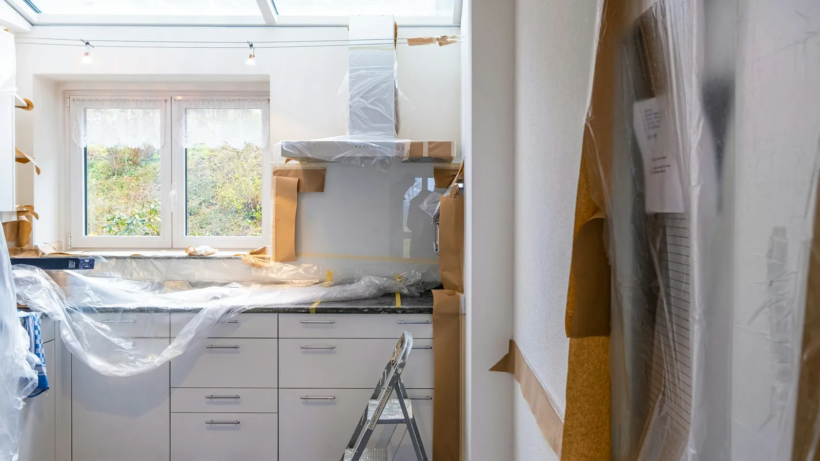 A kitchen in the middle of a remodel, its cabinets and counters wrapped in plastic and paper, a stepladder on the drop cloth