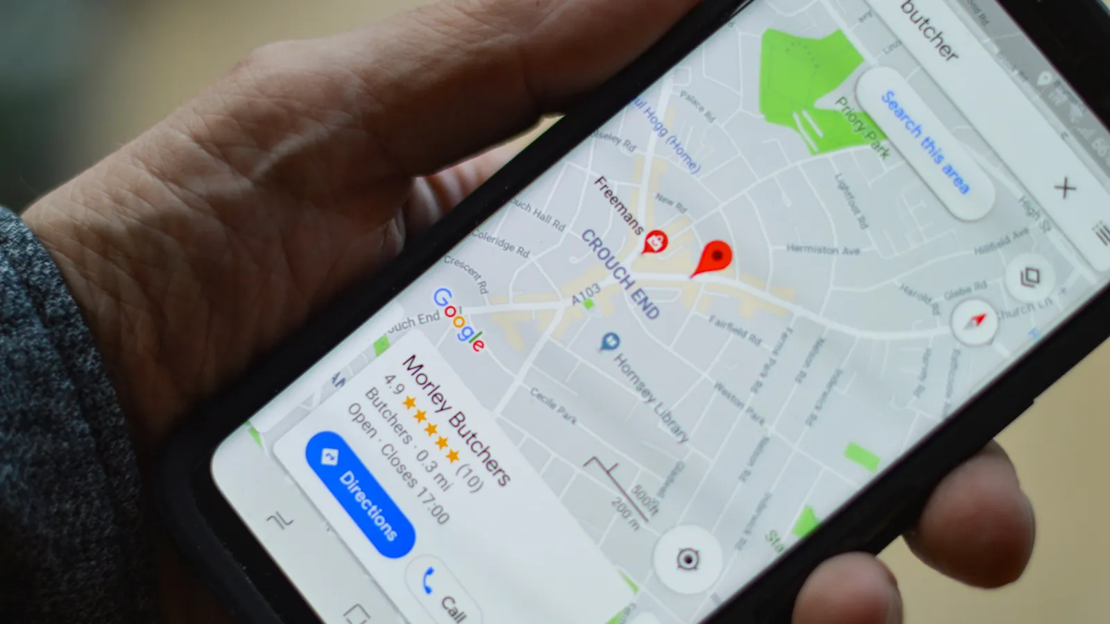 A hand holding a phone with Google Maps open on a search for butchers, a nearby shop's listing showing its rating, hours and a Directions button