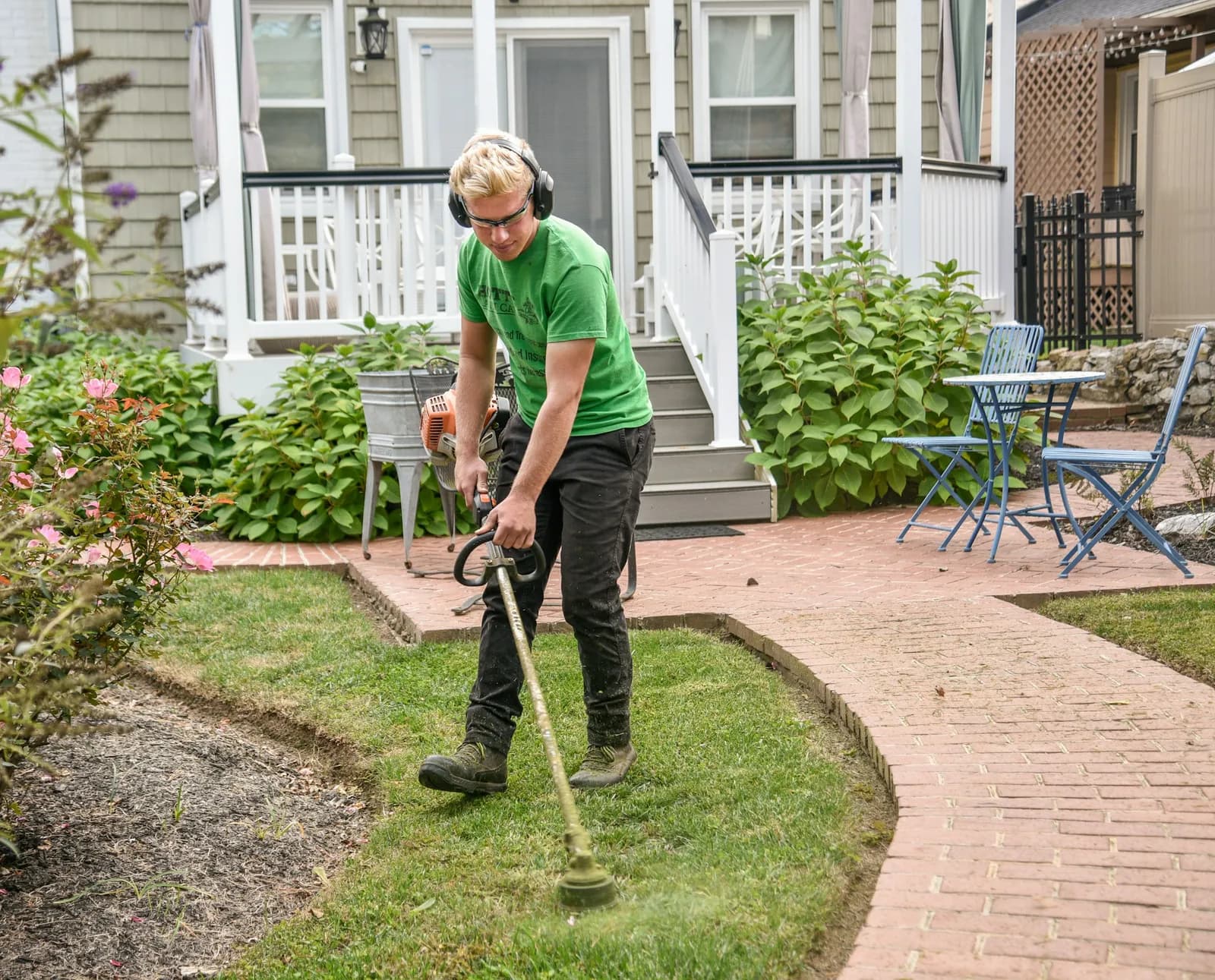 A landscaper in ear protection edging a lawn beside a brick path in front of a house